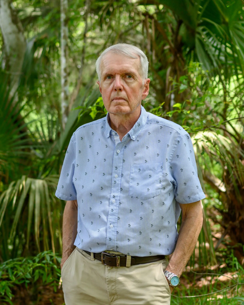 David Swisher, wearing a light blue patterned shirt and khaki pants, stands in front of lush green trees and plants.